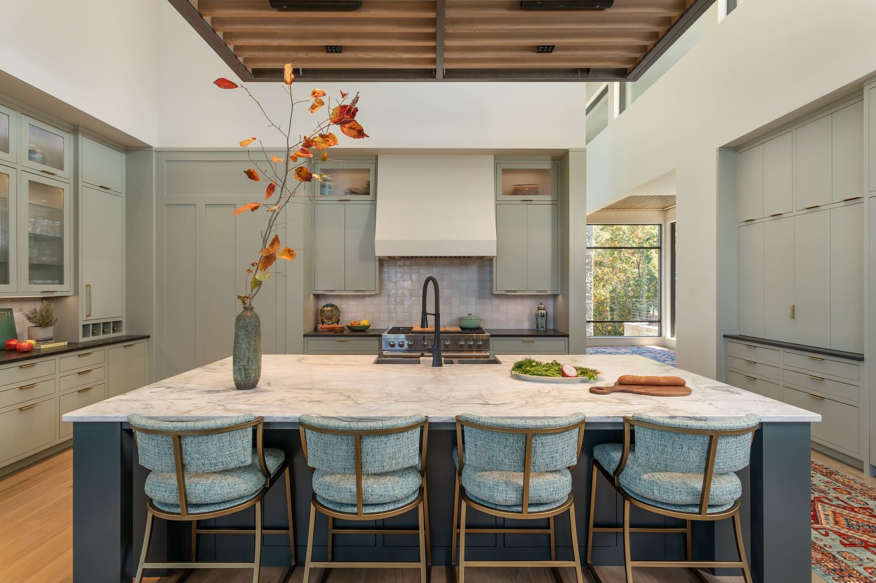 Sage green kitchen with a large marble-topped island, reeded-glass upper cabinets and a plaster range hood